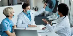 Three healthcare professionals wearing face masks discuss a medical X-ray displayed on a monitor in a clinical setting.