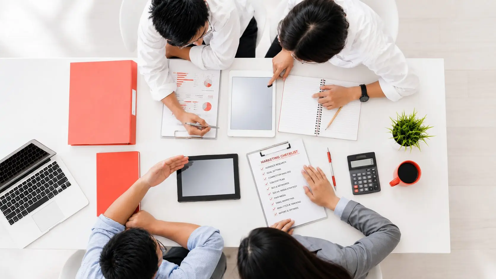 Overhead view of four marketing professionals collaborating at a bright white table with laptops, tablets, reports, a marketing checklist, and coral-red office accessories.