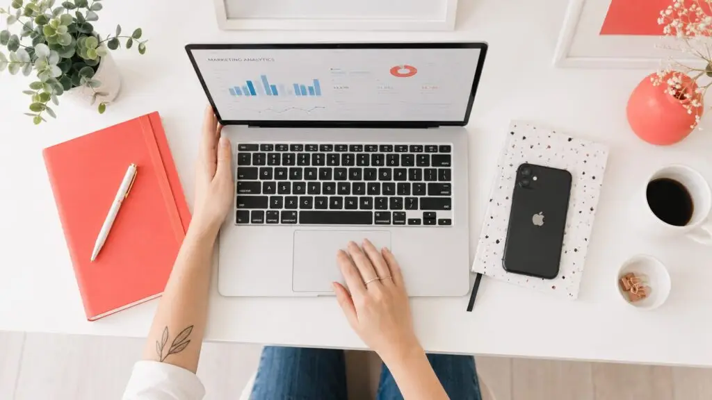 Overhead view of a marketer working on a laptop displaying a marketing analytics dashboard at a bright white desk with coral-red accessories, a phone, coffee, and a small plant.