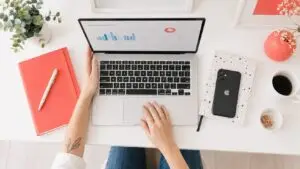 Overhead view of a marketer working on a laptop displaying a marketing analytics dashboard at a bright white desk with coral-red accessories, a phone, coffee, and a small plant.