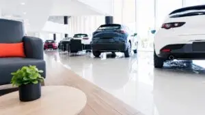 Bright, modern car dealership showroom with white and dark vehicles displayed on glossy floors, black lounge seating with a coral accent pillow, and a small green plant on a light wood table.