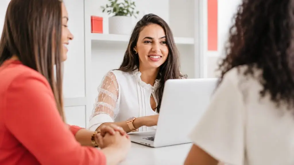 Three women collaborate around a laptop in a bright, modern office with white furnishings and coral-red accents.
