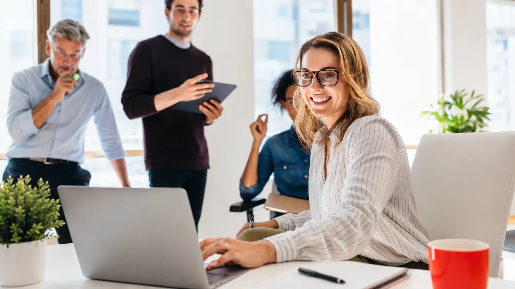Smiling woman working on a laptop in a bright modern office while three colleagues collaborate in the background.