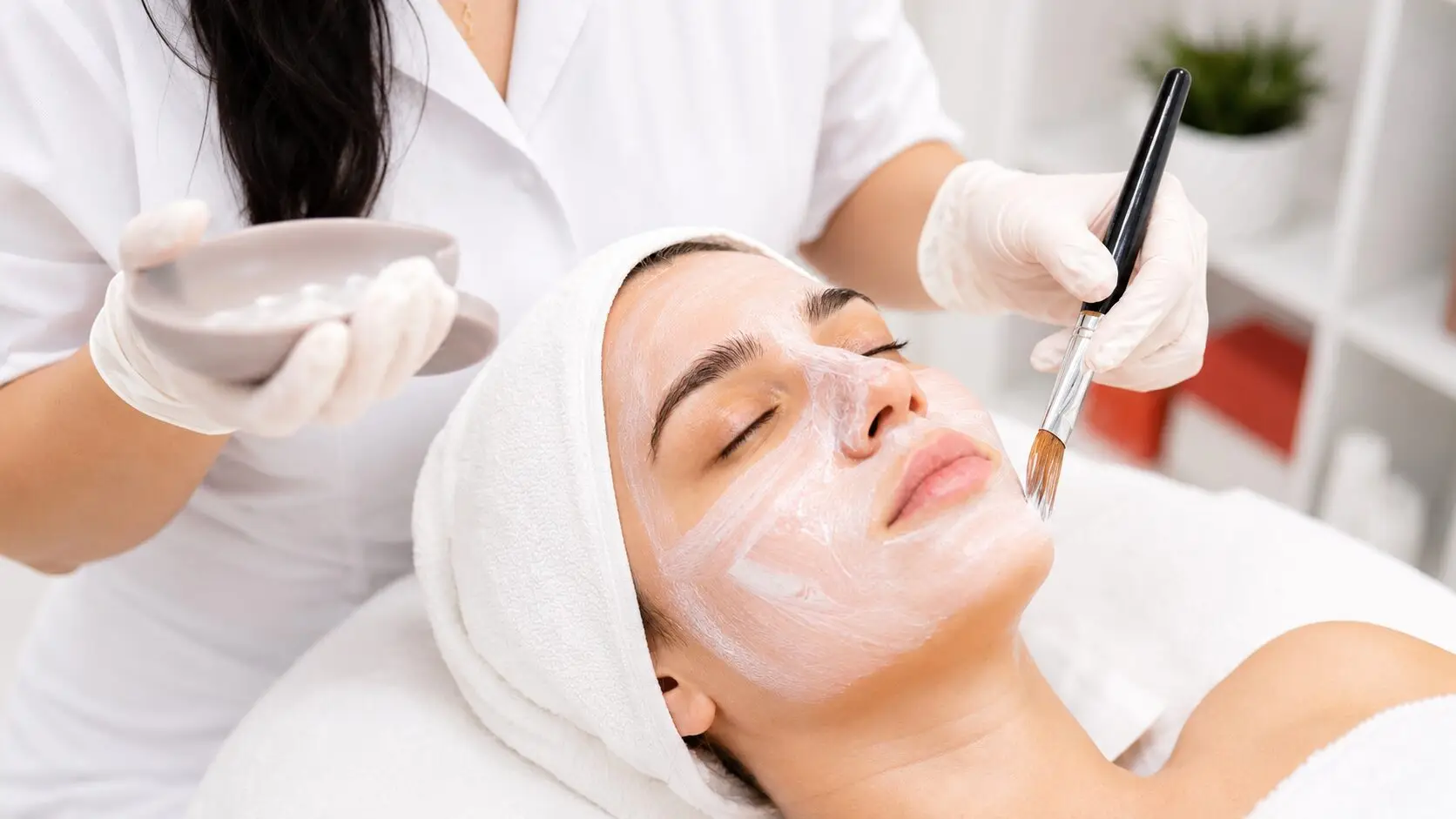 A skincare professional applies a facial mask to a relaxed client in a bright, clean spa treatment room with soft neutral tones and subtle coral accents.