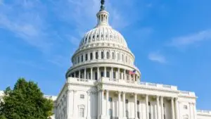 Bright daytime view of the U.S. Capitol building with its white dome, classical columns, American flag, blue sky, and green trees.