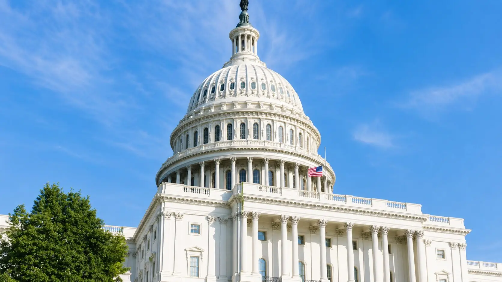 Bright daytime view of the U.S. Capitol building with its white dome, classical columns, American flag, blue sky, and green trees.