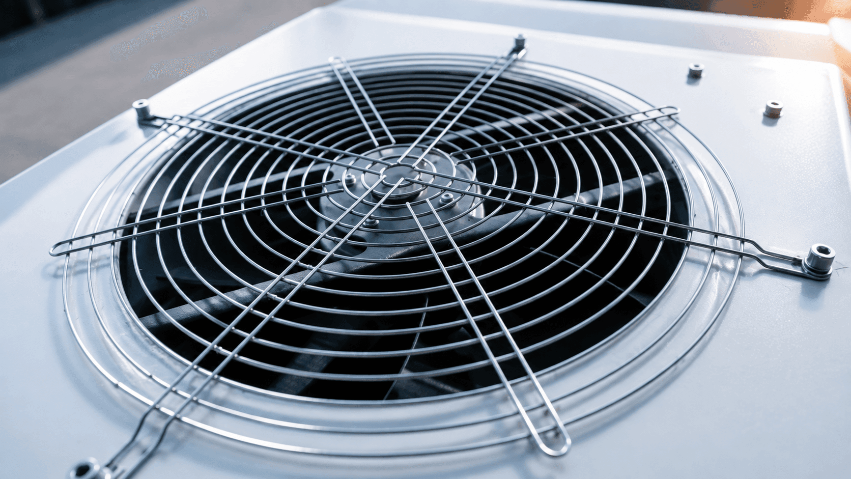 Close-up of an outdoor HVAC condenser fan with a protective metal grille and dark fan blades in bright natural light.