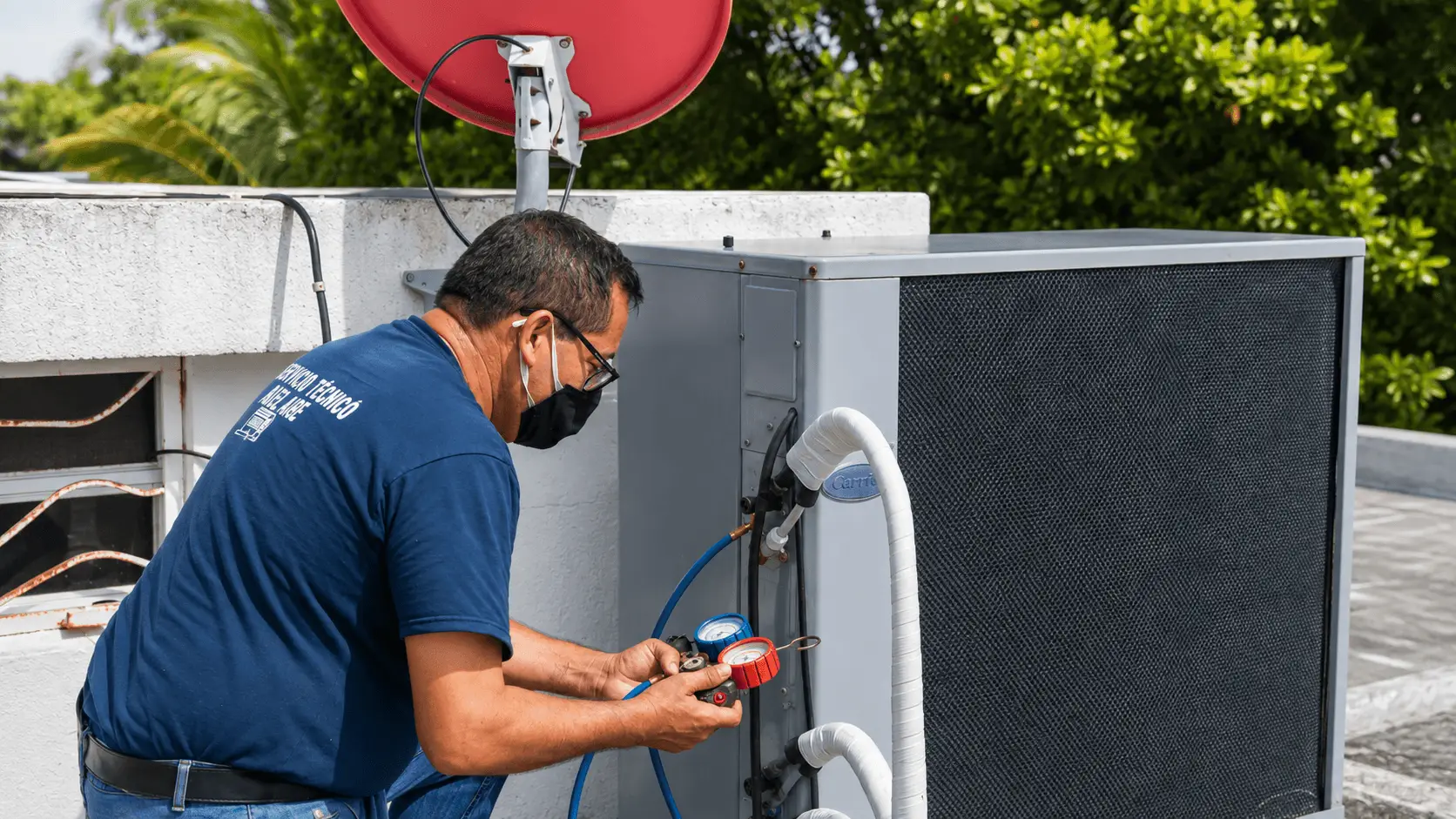 HVAC technician wearing a face mask checks a rooftop air-conditioning unit with red and blue pressure gauges in bright daylight.