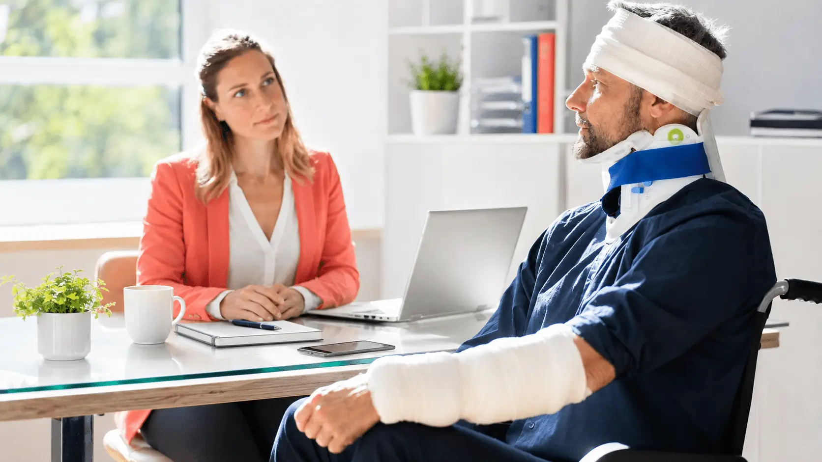 A man wearing a neck brace, head bandage, and arm cast sits in a bright office speaking with a woman in a coral blazer across a desk with a laptop, notebook, phone, coffee cup, and small plant.