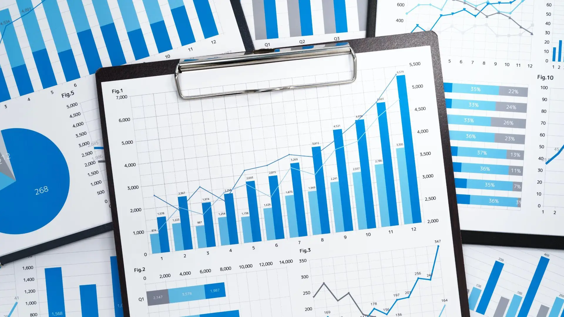 Business analytics dashboard displaying blue bar charts, line graphs, and data visualizations on a tablet surrounded by printed reports.