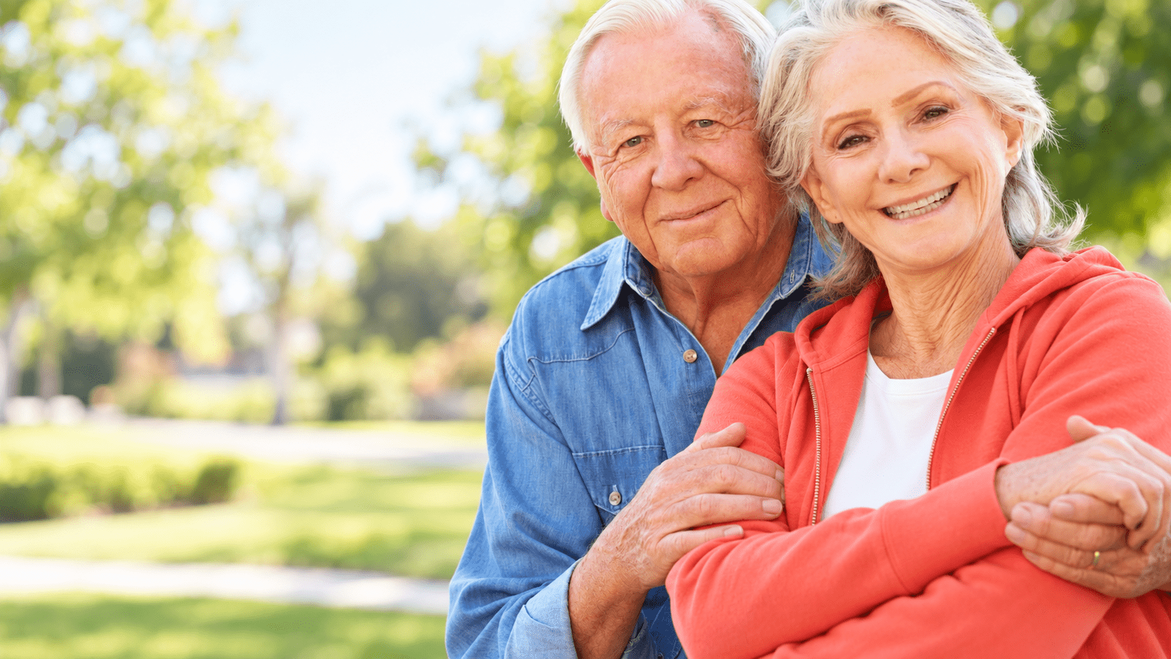 Smiling older couple sitting close together outdoors in a sunny park, with the man’s hand resting gently on the woman’s arm.