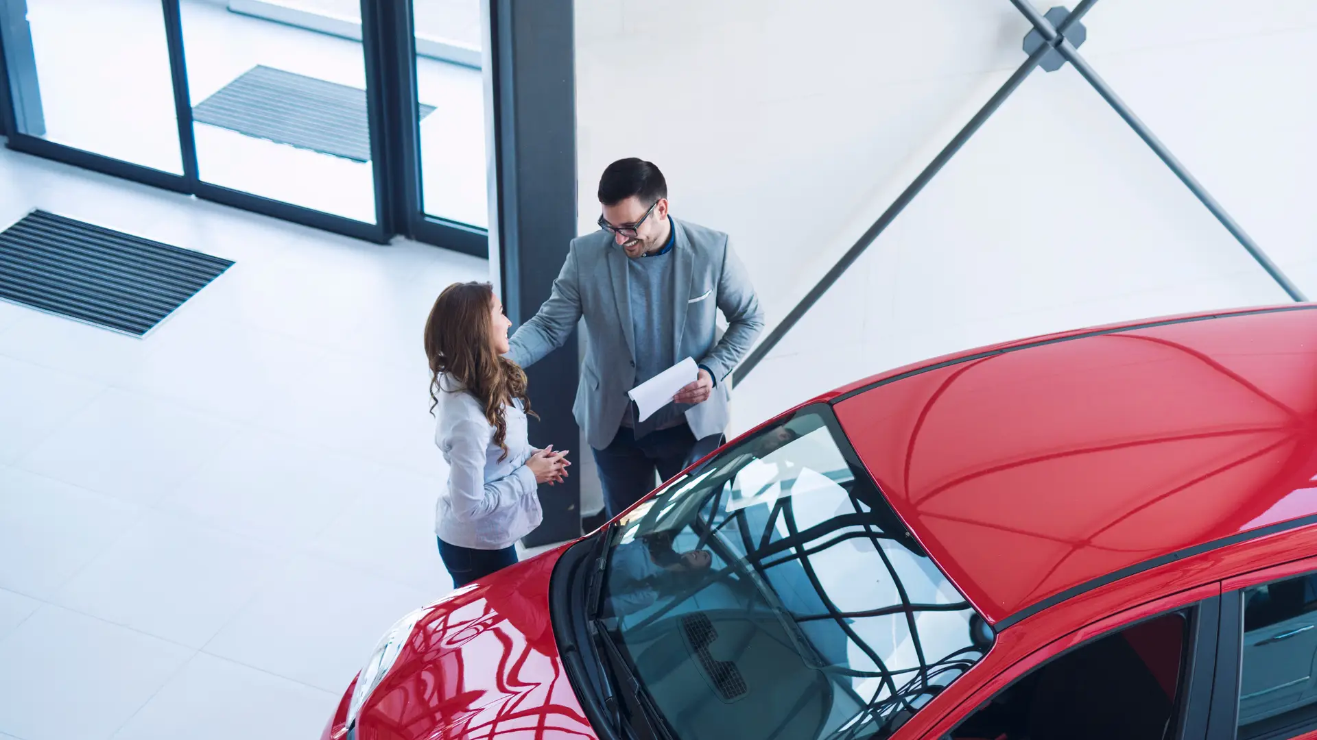 A man holding paperwork talks with a woman beside a red car in a bright showroom.
