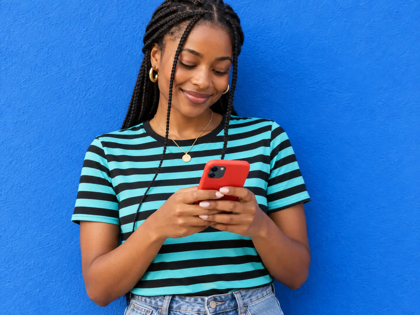 Young Black woman smiling while using a smartphone with a bright Upwynn red case, wearing a turquoise-and-black striped shirt against a vivid blue wall.