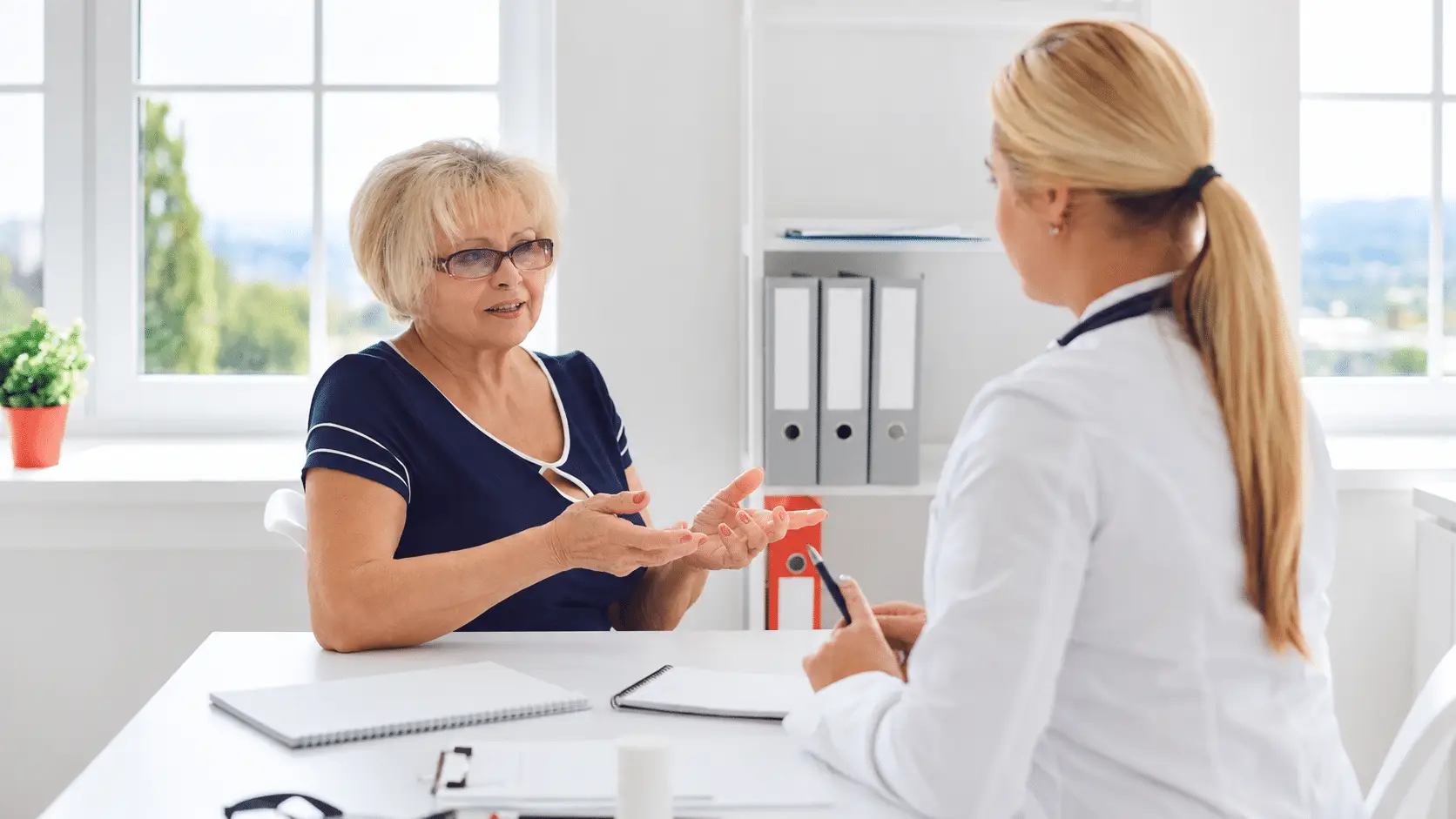 An older woman gestures while speaking with a healthcare professional across a white desk in a bright medical office.