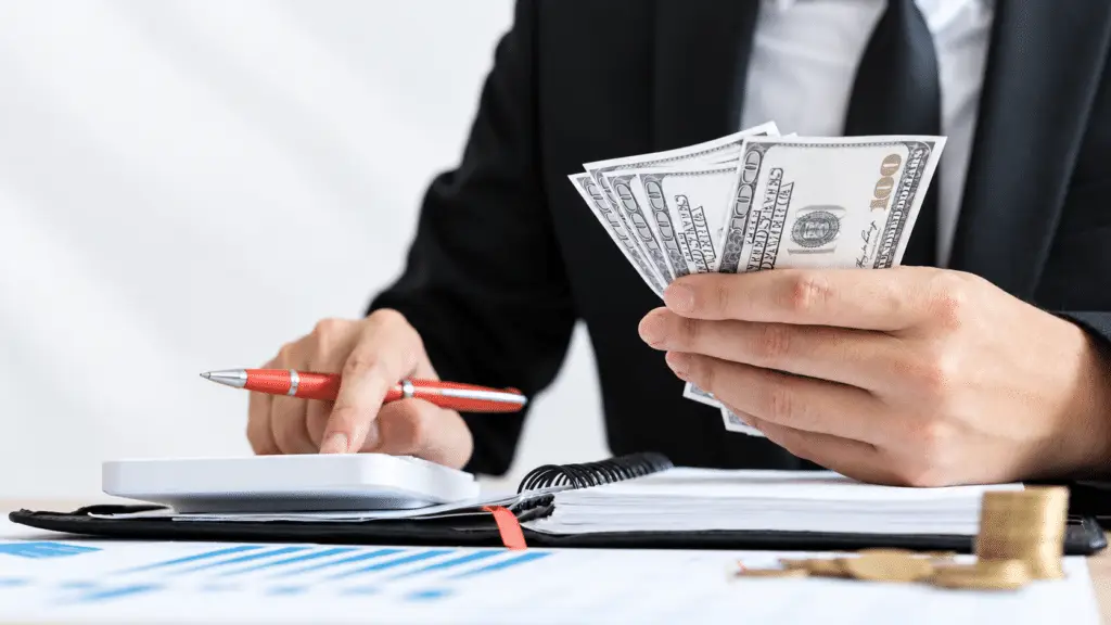 Person in a black suit holding $100 bills while using a calculator, with a coral pen, open notebook, financial charts, and stacked coins on the desk.