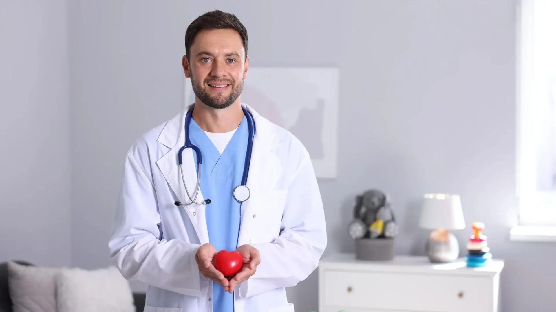 A smiling healthcare professional in a white coat and blue scrubs holds a red heart in both hands in a bright medical office.