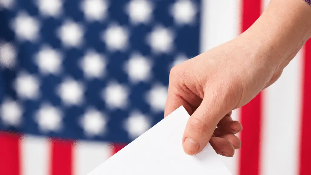 A hand holds a blank white sheet of paper in front of a softly blurred American flag.